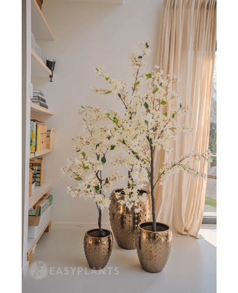 An Artificial Bougainvillea plant 120 cm tall with white blossoms in a textured gold pot stands near a window with sheer curtains, next to shelves full of books and decor in a bright, modern room.