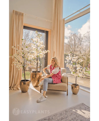 A smiling woman sits in a beige armchair near large windows with transparent curtains, surrounded by Artificial Bougainvillea 120 cm white in gold planters. Sunlight streams in and illuminates the modern, cozy room.