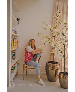 Een persoon zit glimlachend op een houten stoel naast de Kunstplant Bougainvillea 180 cm wit in een goudkleurige pot met textuur. Links staat een boekenplank met boeken en decor; daglicht stroomt naar binnen vanuit een raam aan de rechterkant.