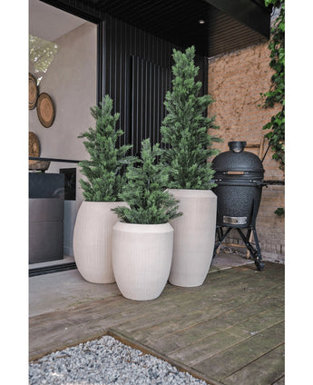 Three high, white planters with Artificial Conifer UV 90cm plant are on a wooden terrace next to a modern building. A black ceramic grill stands nearby against a stone wall lined with vines.