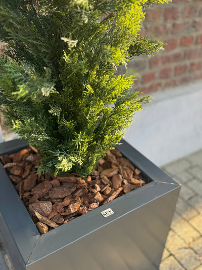 Een close-up van de Kunstplant Conifeer UV 120cm, een realistische groene conifeer in een moderne vierkante grijze plantenbak met bruine schorssnippers, uitgestald tegen een bakstenen muur en betegelde grond.