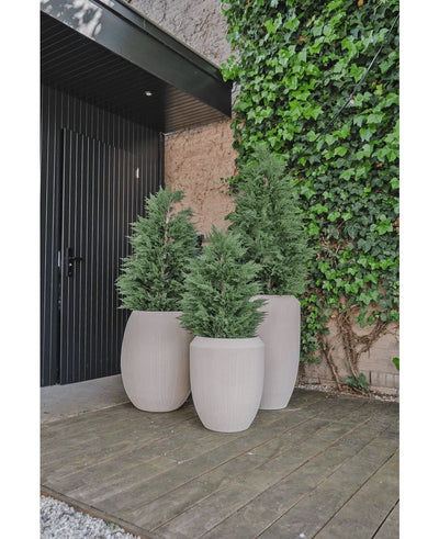 Three high, light-colored planters with luxurious outdoor plants such as Artificial Conifer deluxe UV 100 cm stand on a wooden deck near a wall covered with green ivy, next to a black door and an area with partial gravel.