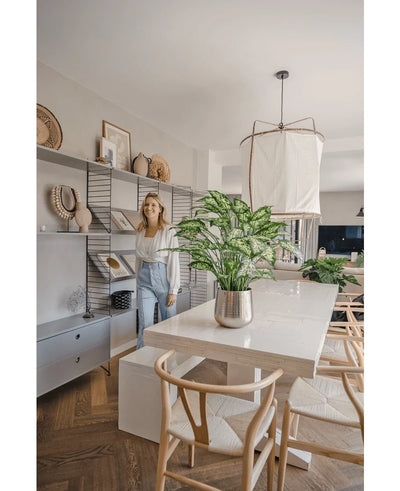 A woman smiles in a bright, modern dining room with wooden chairs, a white table with the Artificial Dieffenbachia 75 cm plant as a centerpiece and open shelves decorated with books and art.
