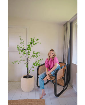 A woman in a pink top and blue jeans sits barefoot on a modern wicker chair next to the Artificial Enkianthus 150 cm plant in a bright, minimalist room with white walls and light carpeting.