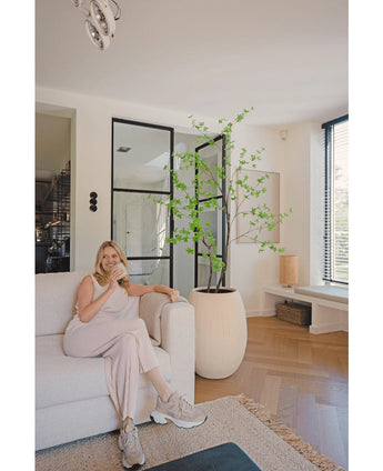 Een vrouw in lichte loungekleding zit op een beige bank en glimlacht met een kopje koffie in een moderne woonkamer met houten vloeren, ramen van vloer tot plafond en de Kunstplant Enkianthus 180 cm als grote potplant.