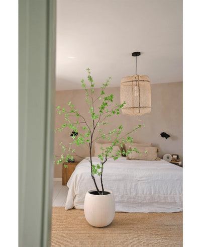 Een minimalistische slaapkamer met de Kunstplant Enkianthus 180 cm als grote potplant, een bed met wit beddengoed, een geweven hanglamp, beige muren en een vloerkleed van natuurvezel voor een eenvoudige en luchtige sfeer.