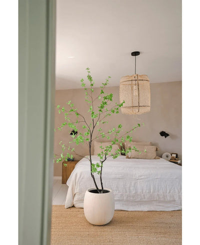 Een minimalistische slaapkamer met beige muren, een wit bed, een natuurlijk geweven tapijt en de Kunstplant Enkianthus 210 cm - een grote potplant met groene bladeren - op de voorgrond. Aan het plafond hangt een geweven hanglamp.