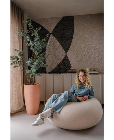 Een vrouw met krullend haar, gekleed in spijkerstof en witte sneakers, glimlacht terwijl ze op een ronde stoel zit. Achter haar staat de Kunstplant Eucalyptus 180 cm, een dressoir en abstracte muurkunst.