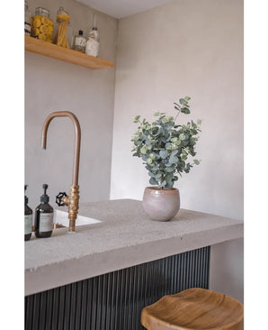 A modern countertop with a bronze tap, soap bottles, a wooden stool and the Artificial Eucalyptus 76 cm. In the background, wooden shelves with jars and bottles are mounted on a light gray wall.