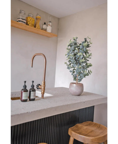 Een modern aanrecht heeft een messing kraan, zeepdispensers, houten krukjes en de Kunstplant Eucalyptus 91 cm. Glazen potten met voorraadartikelen staan op een houten plank tegen een lichtgekleurde muur.