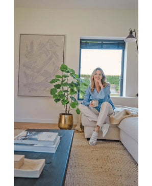 Een vrouw zit op een beige bank in een gezellige, moderne woonkamer, met een mok in de hand en een glimlach. De ruimte wordt gekenmerkt door de Kunstplant Ficus 120 cm in een gouden pot, abstracte kunstwerken en zonlicht dat door blauwe jaloezieën naar binnen schijnt.