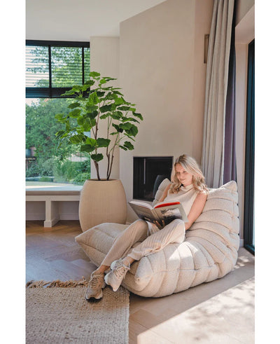 Een vrouw leest in een lichte, moderne woonkamer op een gezellige fauteuil bij het raam, in de buurt van een Kunstplant Ficus 150 cm en zachte gordijnen, omringd door natuurlijk licht.