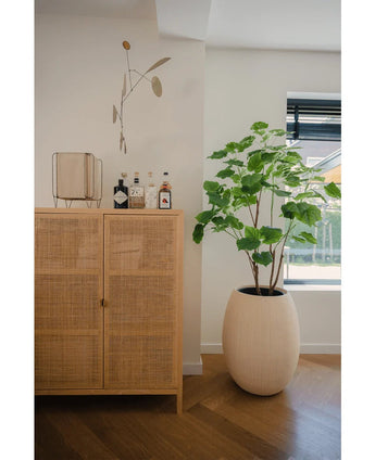 Een lichthouten kast met flessen en decor staat bij een Kunstplant Ficus 150 cm in een crèmekleurige plantenbak bij een raam met jaloezieën, in een lichte moderne kamer met houten vloer.