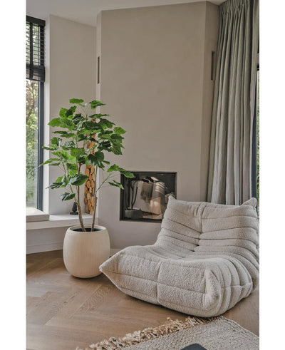 Een gezellige, beige loungestoel met structuur staat naast de Kunstplant Ficus 150 cm in een moderne woonkamer met lichte houten vloeren, neutrale muren, een raam en vloerlange grijze gordijnen.