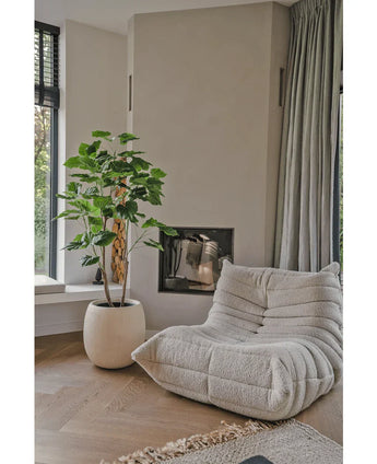 Een gezellige, beige loungestoel met structuur staat naast de Kunstplant Ficus 150 cm in een moderne woonkamer met lichte houten vloeren, neutrale muren, een raam en vloerlange grijze gordijnen.