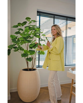 Een vrouw in een gele blazer en witte broek glimlacht terwijl ze de Kunstplant Ficus 150 cm verzorgt in een moderne, goed verlichte kamer met houten vloeren en grote ramen met jaloezieën.