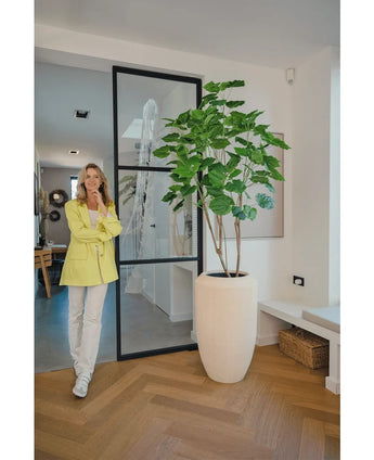 A woman in a yellow blazer and white trousers smiles next to the Artificial Ficus plant 180 cm in a modern, bright room with wooden floor and minimalist decor.