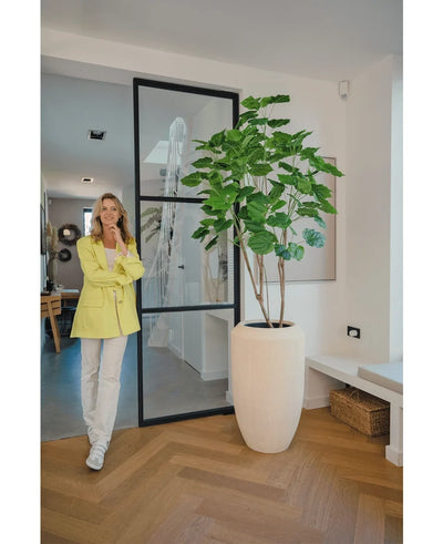 A woman in a yellow blazer and white trousers smiles next to the Artificial Ficus plant 180 cm in a modern, bright room with wooden floor and minimalist decor.