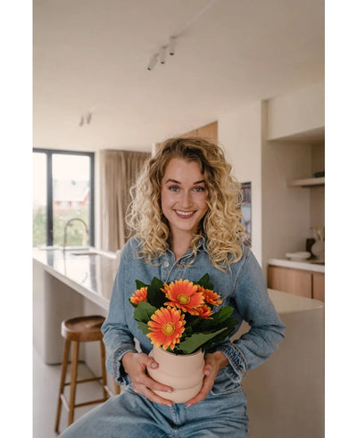 Een glimlachende vrouw met krullend blond haar in spijkerstof zit in een moderne keuken en houdt de Kunstplant Gerbera UV 28 cm oranje vast. Helder natuurlijk licht stroomt door grote ramen achter haar.