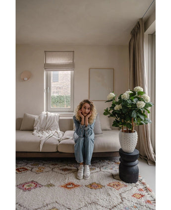 Een vrouw met krullend blond haar zit op een beige bank in een lichte woonkamer en laat haar kin op haar handen rusten. Naast haar staat de Kunstplant Hortensia UV 85 cm groot, verlicht door licht door een raam met beige gordijnen.