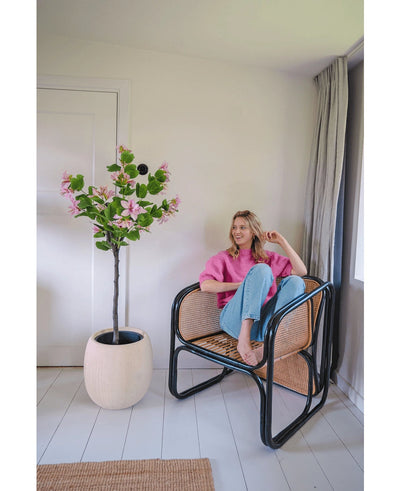 A woman in a pink sweater and blue jeans sits barefoot on a rattan chair next to the Artificial Plant Judas 150 cm, an artificial tree with pink flowers, in a bright, modern room with white walls and floors.