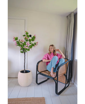 Een vrouw in een roze trui en blauwe spijkerbroek zit op blote voeten op een rotan stoel naast de Kunstplant Judas 150 cm, een kunstboom met roze bloemen, in een lichte, moderne kamer met witte muren en vloeren.