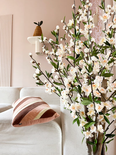 Een gezellige woonkamer met een witte bank, een gestreept roze en bruin kussen en de Kunstplant Kersenbloesem 120 cm crème bloeiende tak op de voorgrond. Een potplant rust op een wandplank tegen een lichtroze muur.
