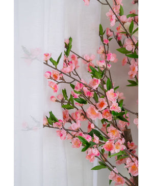 De Kunstplant Kersenbloesem 120 cm roze, met roze kersenbloesem en groene bladeren, staat naast een doorschijnend wit gordijn, de bloemen zacht zichtbaar door de doorschijnende stof.