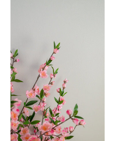 De Kunstplant Kersenbloesem 120 cm roze roze kersenbloesemtakken met groene bladeren die linksonder uitsteken op een effen, lichtbeige achtergrond.
