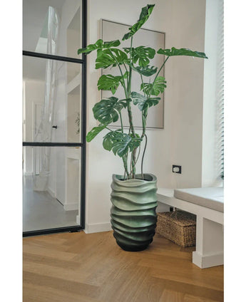 The Artificial Plant Monstera 150 cm, with its large green leaves, stands in a pot with a wavy pattern on a wooden floor in a modern, bright room with a glass door, framed art and a wicker basket nearby.