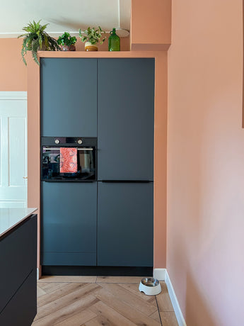 A modern kitchen corner with matte dark cabinets, a built-in oven and a light wooden floor. The Artificial Plant Monstera 18 cm and a green glass bottle are on top of the cabinets; a pet food bowl sits on the peach wall.