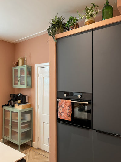 Een moderne keuken met matte donkere kasten, een ingebouwde oven en een handdoek met patroon. Bovenop de kasten staan potplanten zoals Kunstplant Monstera 18 cm en glazen flessen, terwijl een mintgroene kast in de hoek koffiebenodigdheden bevat.