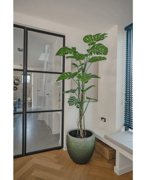 De Kunstplant Monstera 180 cm, een hoge potplant met grote gespleten bladeren, staat in een moderne, lichte kamer naast een glazen deur, houten vloer, bank en blauwe verticale lamellen.