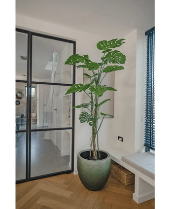 De Kunstplant Monstera 180 cm, een hoge potplant met grote gespleten bladeren, staat in een moderne, lichte kamer naast een glazen deur, houten vloer, bank en blauwe verticale lamellen.