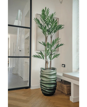 De Kunstplant Parlour Deluxe 120 cm heeft dunne stelen en lange groene bladeren in een golvende groene vaas, perfect voor lichte moderne kamers met lichte houten vloeren en grote ramen.