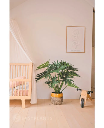 A cozy, minimalist children's room with a wooden cradle and canopy on the left, an Artificial Philodendron UV 90 cm plant in the middle as a showpiece, a wooden toy bicycle on the right and a simple line art print above it.