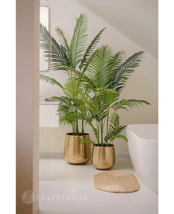 Twee Kunstplant Phoenix 160 cm met lange, groene bladeren in glanzende gouden potten geven een luxe touch aan de witte badkamervloer bij het bad en de ronde geweven mat. Op de achtergrond zijn een spiegel en een wit aanrecht te zien.