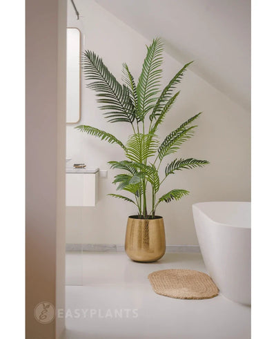 The Artificial Plant Phoenix 180 cm in a gold pot stands next to a white bath and sink in a modern minimalist bathroom with white walls and floor. Nearby is a round woven mat on the floor.