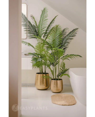An Artificial Plant Phoenix 180 cm with lush green leaves in a shiny gold pot stands next to a white bath and a beige woven mat in a modern, minimalist bathroom, complemented by a mirror and a window for a serene look.