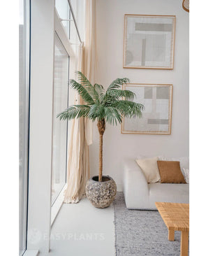 De Kunstplant Phoenix Palm De Luxe 180 cm in een pot met structuur staat bij grote ramen in een lichte woonkamer met neutrale inrichting, met een witte bank, beige kussens, een houten tafel en abstracte kunst aan de muur.