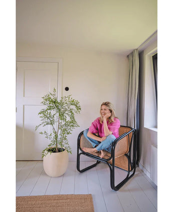 A woman in a pink shirt and jeans sits barefoot on a woven chair, smiling and relaxing in a bright, minimalist room with white walls and floors. Next to her is the Artificial Ruscus 120 cm plant.