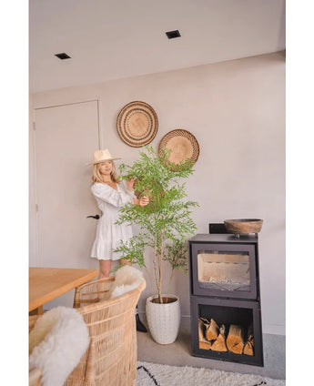 A woman in a white dress and hat stands next to the Artificial Ruscus plant 150 cm, which adds a natural flair to a modern room with wicker furniture, wall baskets and a black wood-burning stove - perfect for decorative interiors.