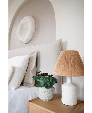 Een moderne slaapkamer met een neutraal kleurenpalet heeft een bed met wit beddengoed, een houten nachtkastje, een lamp van gestructureerd keramiek met geweven kap, de Kunstplant Saxifraga 27 cm, en een ronde witte wandsculptuur boven het hoofdeinde.