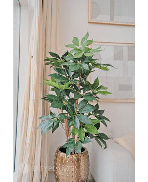 De Kunstplant Schefflera 150 cm met brede groene bladeren staat in een goudkleurige pot naast een crèmekleurige bank en lichte gordijnen, terwijl aan de muur abstracte kunst hangt.