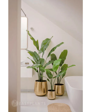 Een Kunstplant Strelitzia Nicolai Deluxe 165 cm in een glanzende gouden pot staat op de vloer naast een wit bad in een moderne, minimalistische badkamer met lichte muren en een kleine spiegel boven een witte wastafel.