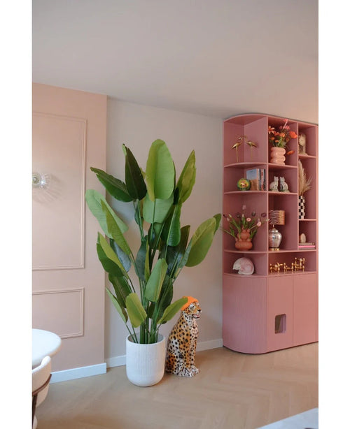 The Artificial Plant Strelitzia Real Touch Deluxe 210 cm stands next to a ceramic cheetah figure with an orange hat, next to a high pink shelf full of vases, flowers and ornaments in a pastel-colored room.