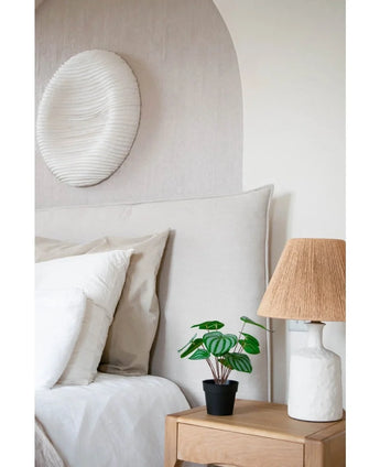 A cozy bedroom has a bed with neutral bedding, a light wooden bedside table with the Artificial Watermelon plant 29 cm and a white lamp with a wicker shade. A white textured wall sculpture hangs above the bed.