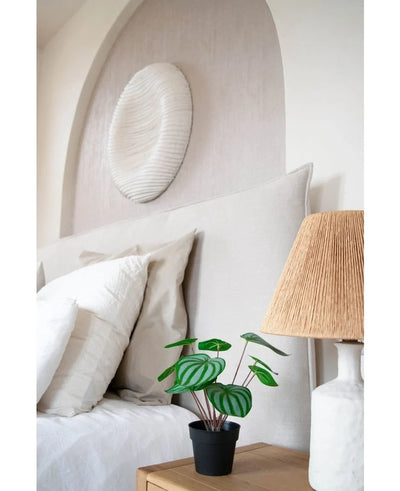 A neatly made bed with white pillows and bedding stands next to a wooden bedside table with the Artificial Watermelon plant 29 cm and a lamp with a woven shade; above the headboard hangs a white textured wall sculpture.