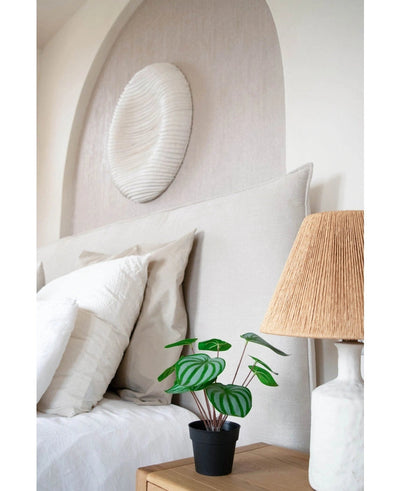 Een keurig opgemaakt bed met witte kussens en beddengoed staat naast een houten nachtkastje met de Kunstplant Watermeloen 29 cm en een lamp met een geweven kap; boven het hoofdeinde hangt een wandsculptuur met witte textuur.