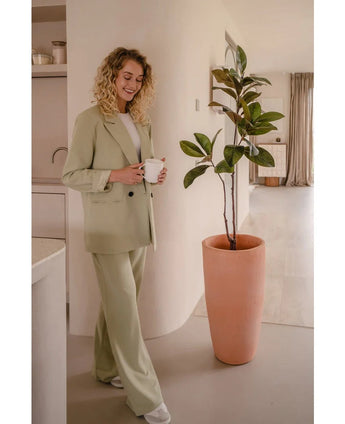 Een vrouw met krullend blond haar, gekleed in een lichtgroen pak en wit topje, staat binnenshuis te glimlachen met een mok naast een Kunstplant Yulaniaboom 120 cm in een moderne, neutraal getinte kamer.